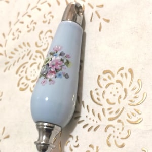 Op de afbeelding: Een lichtblauwe flesstopper met een bloemenontwerp in roze, blauw en wit. De stopper heeft een zilverkleurige metalen boven- en onderkant. Het bloemenontwerp staat op de voorkant van de stopper.