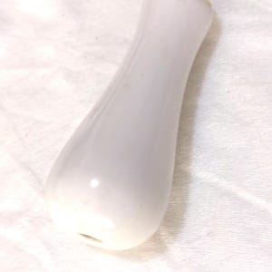 May include: A white, elongated ceramic object with a smooth, glossy finish. The shape is slightly curved, wider at the base and tapering towards the top. The object has a small opening at the bottom.