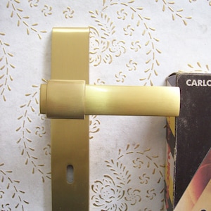 May include: A gold door handle with a square backplate and a lever handle. The handle is mounted on a white wall with a floral pattern.