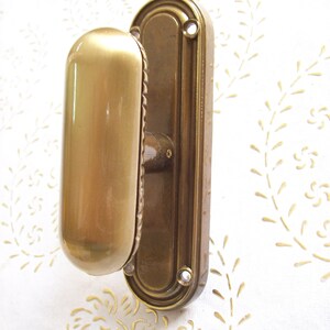 Könnte beinhalten: Ein Türgriff aus Messing im Vintage-Stil mit einem langen, abgerundeten Griff und einer ovalen Rückplatte. Der Griff hat eine glatte, polierte Oberfläche, und die Rückplatte weist dekorative Details auf. Montiert an einer weißen Wand mit floralem Muster.