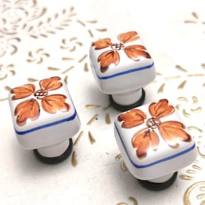 Puede incluir: Tres pomos de armario cuadrados blancos con un diseño floral naranja pintado a mano. Cada pomo tiene un borde azul y una base negra. Los pomos están colocados sobre un fondo blanco con un patrón floral dorado.