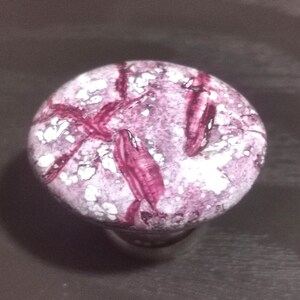 May include: A round cabinet knob featuring a marbled pattern in shades of dusty rose, deep magenta, and white. The surface has a glossy, smooth finish with irregular, vein-like streaks of dark pink running across the circular top face.