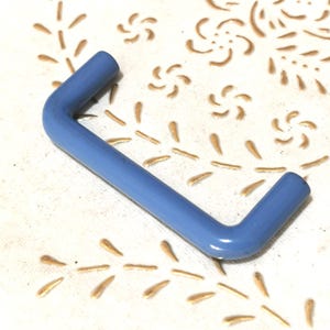 Könnte beinhalten: Ein hellblauer, L-förmiger Metallgriff. Der Griff befindet sich auf einer weißen Oberfläche mit einem goldenen Blumenmuster. Der Griff ist etwa 10 cm lang und 5 cm breit.