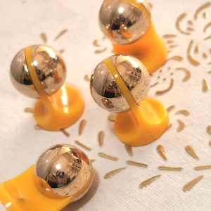 May include: Four decorative silver and yellow magnets. Each magnet features a shiny silver sphere with a yellow band, mounted on a bright yellow base. The magnets are arranged on a white surface with gold-colored decorative accents.