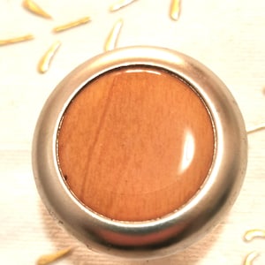 Puede incluir: Un pomo redondo de madera con un marco de metal plateado. La madera tiene un color marrón claro con un sutil patrón de veta.