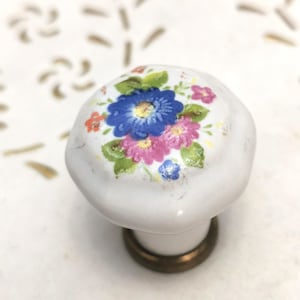 Puede incluir: Pomo de armario de cerámica blanca con un diseño floral con flores azules, rosas y naranjas. El pomo tiene una base de latón y está sobre un fondo blanco con detalles dorados. Un artículo de ferretería decorativo.