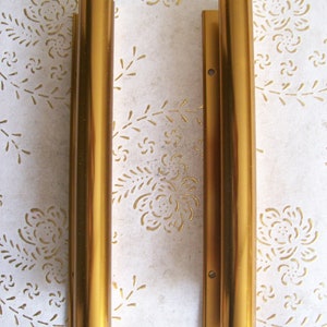 Könnte beinhalten: Zwei goldfarbene Metall-Schubladengriffe, jeweils mit zylindrischer Form und einem leicht unterschiedlichen Design. Die Griffe sind etwa 25 cm lang.