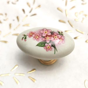 May include: White ceramic cabinet knob with a floral design featuring pink flowers and green leaves. The knob has a gold metal base.