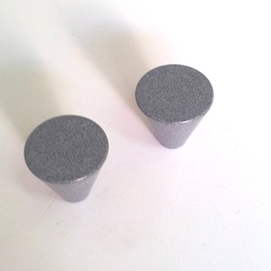 Könnte beinhalten: Zwei graue, kegelförmige Schrankknöpfe mit strukturierter Oberfläche. Die Knöpfe haben eine flache, kreisförmige Oberseite und eine konische Basis. Die Knöpfe bestehen aus einem festen Material und eignen sich für Schubladen oder Schränke.