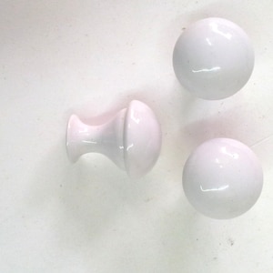 Op de afbeelding: Drie witte keramische kastknoppen. Twee zijn rond en één is rond met een kleine steel.