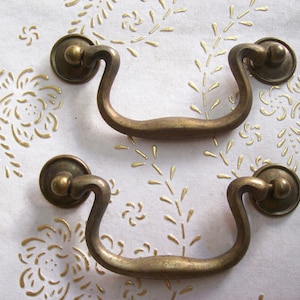 May include: Two antique brass drawer pulls with a curved handle design. Each pull has a circular base and a curved handle, showcasing a vintage aesthetic. The pulls are set against a white background with gold floral patterns.