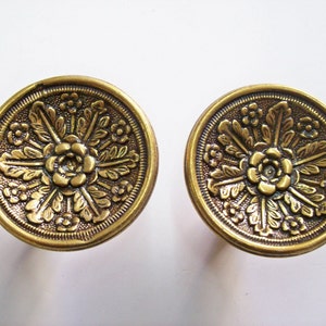 Puede incluir: Dos lazos para cortinas de metal dorado con un diseño floral. Los lazos son redondos y tienen un diseño en relieve de una flor con hojas.