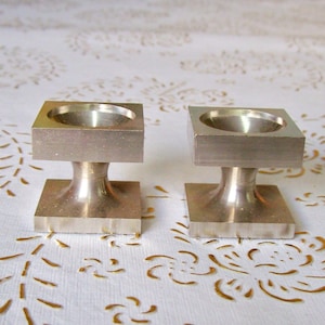 Può includere: Due porta uova in metallo argentato con base quadrata e parte superiore rotonda. I porta uova sono su una tovaglia bianca con un motivo dorato.