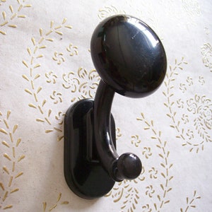 May include: A black wall hook with a rounded top and a curved hook, mounted on a rectangular base. The background is a cream-coloured wall with a gold floral pattern. The hook is a classic design.