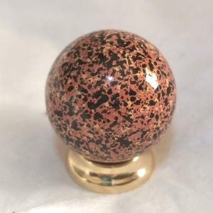 Peut inclure: Un bouton de meuble sphérique présentant un motif tacheté de points noirs et fauves sur un fond rose poudré, monté sur une base métallique circulaire dorée polie. La surface de la sphère possède une finition lisse, brillante et réfléchissante.