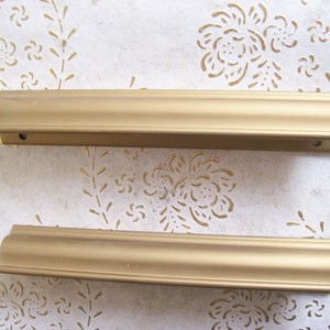 May include: Two gold-coloured metal drawer pulls with a decorative edge. The pulls are approximately 30 cm long.