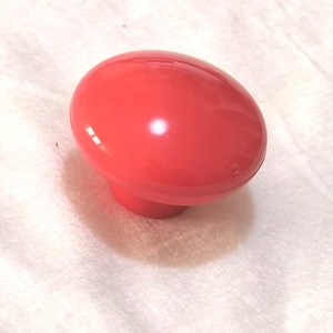 May include: A bright red, oval-shaped cabinet knob with a rounded base. The knob is glossy and reflects light, suggesting a smooth finish. The knob is likely made of plastic or a similar material. This hardware is suitable for drawers or cabinets.