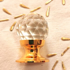 Op de afbeelding: Een heldere glazen kristallen kastknop met een gouden metalen basis. De knop heeft een facetgeslepen ontwerp en is perfect om elke kamer een vleugje elegantie te geven.