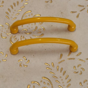 Op de afbeelding: Twee gele metalen kastgrepen met een afgeronde vorm. De grepen bevinden zich op een wit oppervlak met een gouden bloemenpatroon.