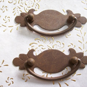 May include: Two antique-style, rusty metal drawer pulls with ornate details. Each pull has a curved handle and a rectangular plate.