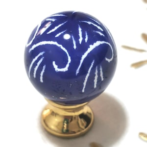 Puede incluir: Pomo decorativo para armario con una esfera de cerámica azul cobalto adornada con patrones blancos en espiral. La esfera se asienta sobre una base de metal dorado, añadiendo un toque de elegancia a los muebles. Diámetro aproximado de 3 cm.