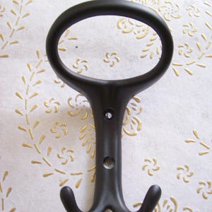 Puede incluir: Un gancho de pared de metal marrón oscuro con una parte superior ovalada y dos ganchos inferiores. El gancho tiene un diseño decorativo y dos orificios para tornillos para el montaje. Fondo blanco con patrón floral dorado.