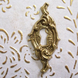 Op de afbeelding: Een goudkleurige sleutelgatbedekking met een sierlijk ontwerp. De afdekking is gemaakt van metaal en heeft een decoratief, wervelend patroon.