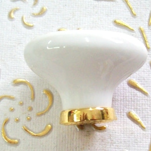 May include: White ceramic knob with a gold rim. The knob is round and has a slight indentation in the center. It is a decorative knob that could be used for cabinets, drawers, or doors.