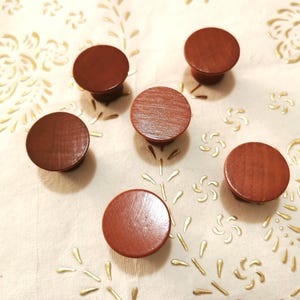 May include: Six round wooden knobs in a reddish-brown color. The knobs are arranged in a semi-circle on a white surface with a gold floral pattern.