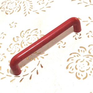 May include: A red, rectangular handle with curved ends. The handle is placed on a white surface decorated with a gold floral design. This handle is likely for a cabinet or drawer, or similar furniture.