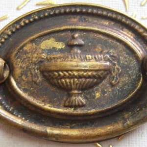 Puede incluir: Tirador de armario de latón antiguo con una placa ovalada y un diseño decorativo de una cesta con un motivo floral.