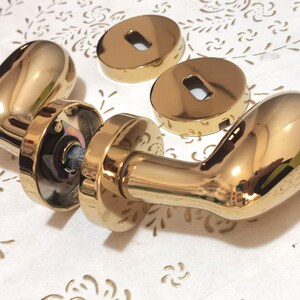 May include: Gold-toned door hardware set, including two door handles and two keyhole covers. The handles have a curved design and a shiny finish. The keyhole covers are round with a rectangular opening. The hardware is likely for interior doors.