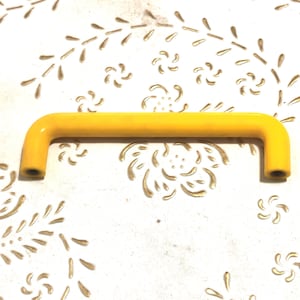 Op de afbeelding: Een heldergele kastgreep met een gebogen ontwerp. De handgreep heeft een effen kleur en een cilindrische vorm. De achtergrond is een wit oppervlak met een gouden bloemenpatroon.