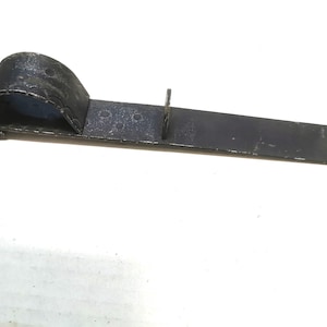 May include: A dark gray metal tool with a curved handle and a flat, rectangular body. The handle has a decorative, scalloped edge. The tool appears to be made of iron or steel and is likely used for a specific mechanical purpose.