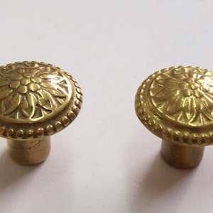 Puede incluir: Dos pomos de metal dorado con un diseño floral. Los pomos tienen una parte superior redonda y una base cilíndrica.