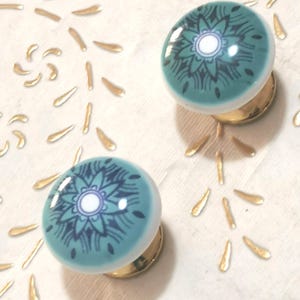 May include: Two teal and white ceramic cabinet knobs with a floral design in dark teal. The knobs have a gold-colored base and are set against a white background with gold leaf accents. These knobs are perfect for adding a touch of elegance to any furniture.