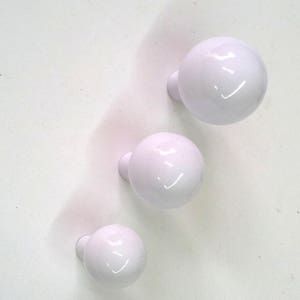 Può includere: Tre pomelli in ceramica bianca di forma rotonda con un piccolo gambo sul retro. I pomelli sono disposti verticalmente, con il pomello più grande in alto e il pomello più piccolo in basso.
