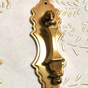 May include: A gold-toned door knocker with an ornate design. The knocker is shaped like a bell with a rounded top and a pointed bottom. It has a decorative handle that is also gold-toned.