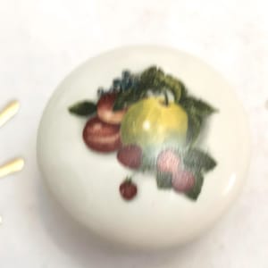 Op de afbeelding: Een witte, ronde keramische knop met een fruitontwerp. Het ontwerp bevat een groene appel, rode en roze bessen en groene bladeren. Deze decoratieve knop is geschikt voor kasten of lades.