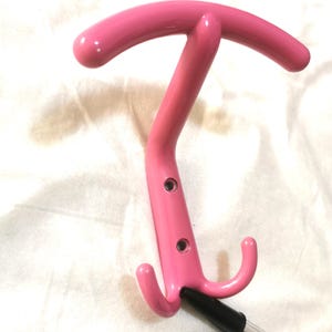 May include: A bright pink metal coat rack with a T-shaped top and two curved hooks. The rack has a glossy finish and two small holes. The rack is designed for hanging coats, hats, or other items.