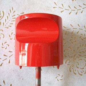 May include: A red plastic knob with a screw thread and three small feet on the bottom. The knob is attached to a metal rod.