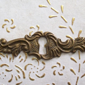 May include: Ornate brass keyhole cover with a decorative design. The cover is a light gold color and has a keyhole in the center.