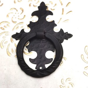 Op de afbeelding: Een zwarte, sierlijke trekker met een ringhandvat. De decoratieve hardware heeft een getextureerd oppervlak en een bloemenontwerp. De achtergrond is wit met goudkleurige bloemaccenten.