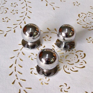 Könnte beinhalten: Drei runde silberne Schrankknöpfe mit glänzender Oberfläche. Die Knöpfe befinden sich auf einer weißen Oberfläche mit einem goldenen Blumenmuster.