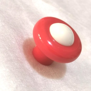 May include: A red mushroom-shaped cabinet knob with a white centre. The knob has a rounded top and a short, cylindrical base. The knob is made of a glossy material and is set against a soft, white background.