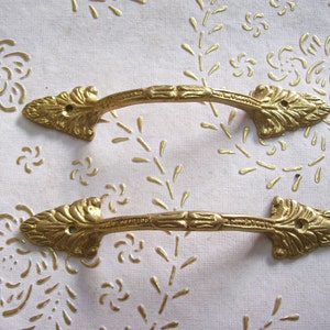 Puede incluir: Dos tiradores de puerta de armario de metal dorado con extremos en forma de hoja adornados. Los tiradores son curvos y tienen un diseño decorativo en el centro.