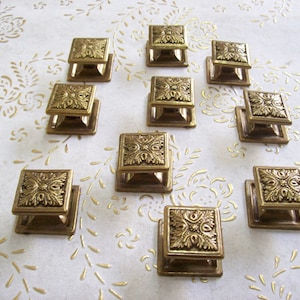 Op de afbeelding: Tien gouden kastknoppen met sierlijke bloemmotieven. Elke knop is vierkant met een reliëfmotief en een kleine basis.