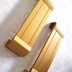 Könnte beinhalten: Zwei goldfarbene Wanddekorationen aus Metall mit vertikalem Lamellen-Design. Die Stücke sind an einer weißen Wand mit einem Blumenmuster angebracht.