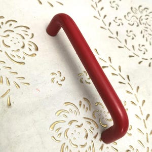 Puede incluir: Un tirador rojo curvado sobre una superficie blanca con recortes florales dorados. El tirador es de un rojo vibrante y la superficie tiene un aspecto envejecido. El diseño floral añade un elemento decorativo.