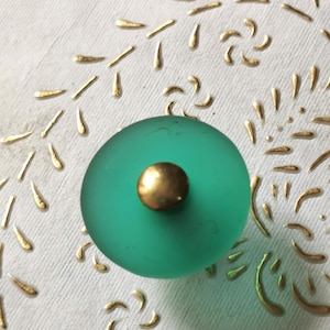 Può includere: Un pomello rotondo in vetro verde turchese con un centro in metallo dorato. Il pomello è su una superficie bianca con un motivo dorato.